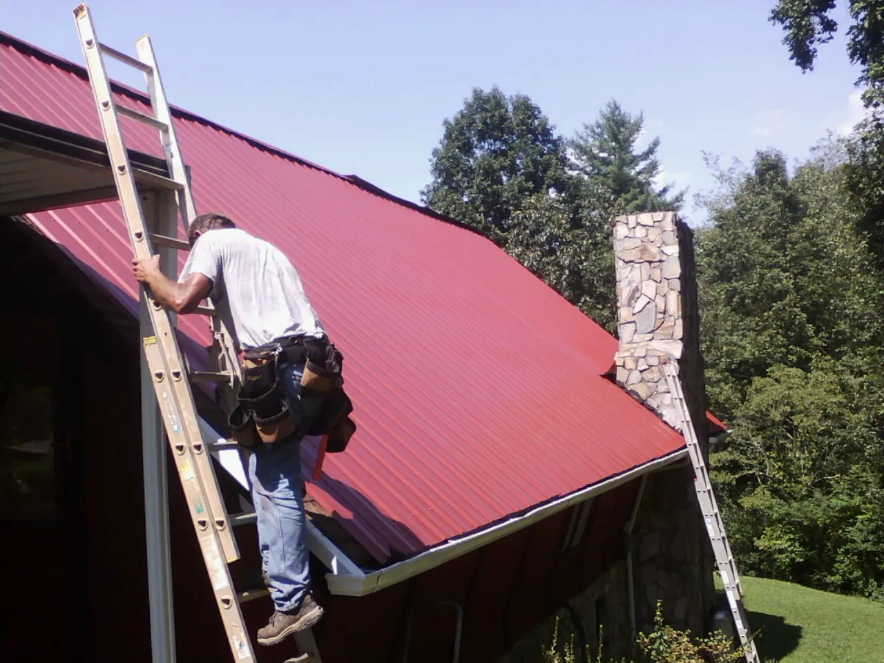 Roofer on a ladder beside a red metal roof and stone chimney — Soffit & Fascia Repair by Roofing Brothers in Snowmass